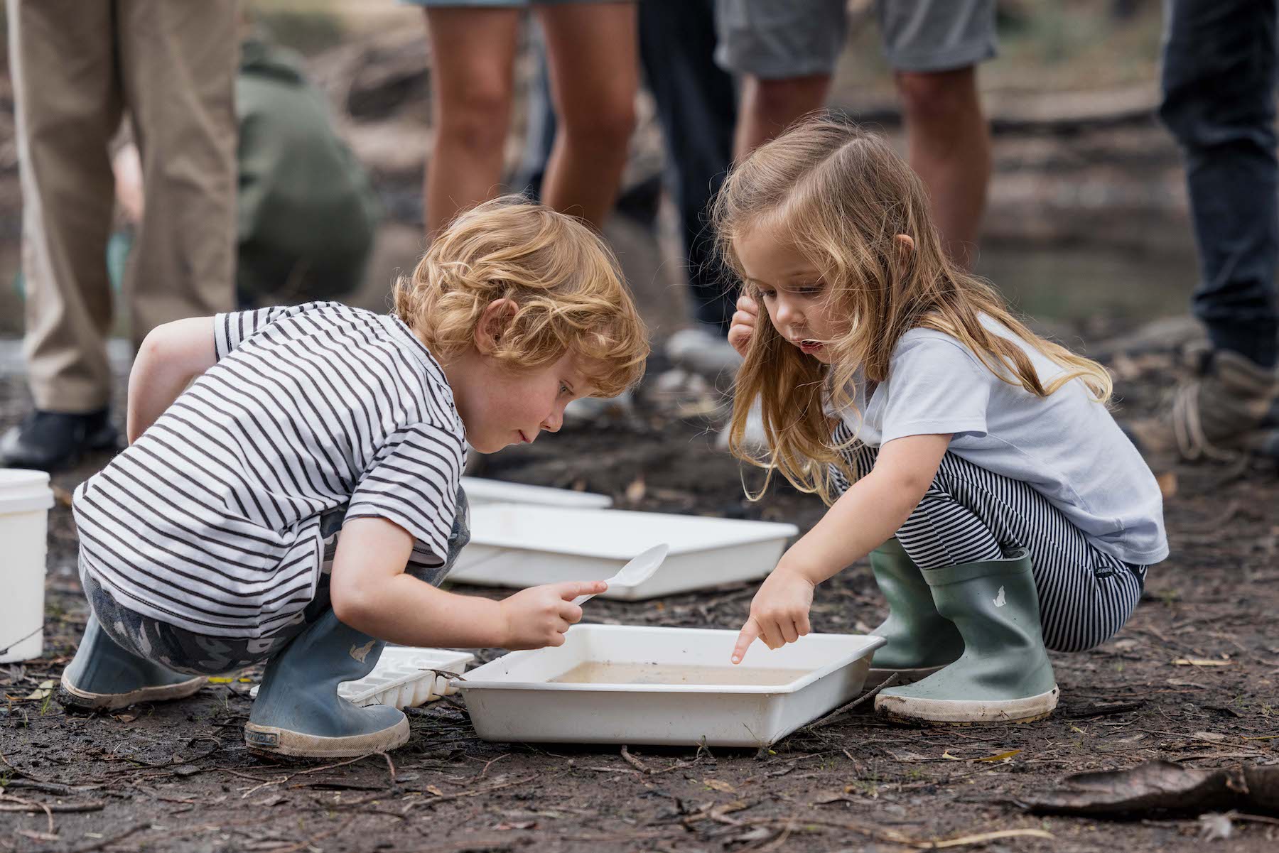 Two young children looking with interest at the contents of a water-filled plastic tray placed on the ground outside.