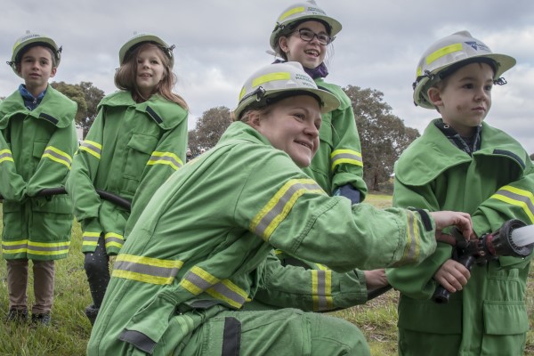 A group of children and a firefighter all dressed in firefighting uniform, using a fire hose.