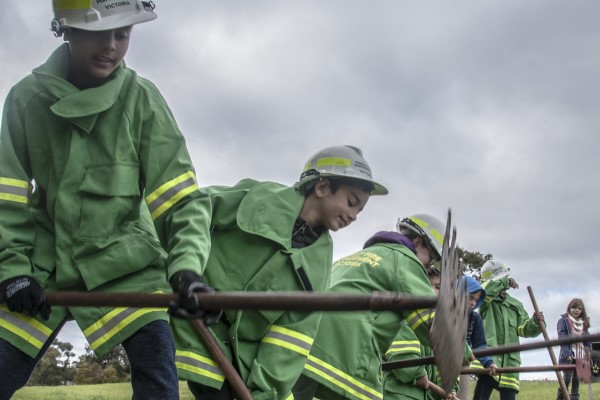 A group of children dressed in firefighting overalls, using rakehoes to clear a fire break.